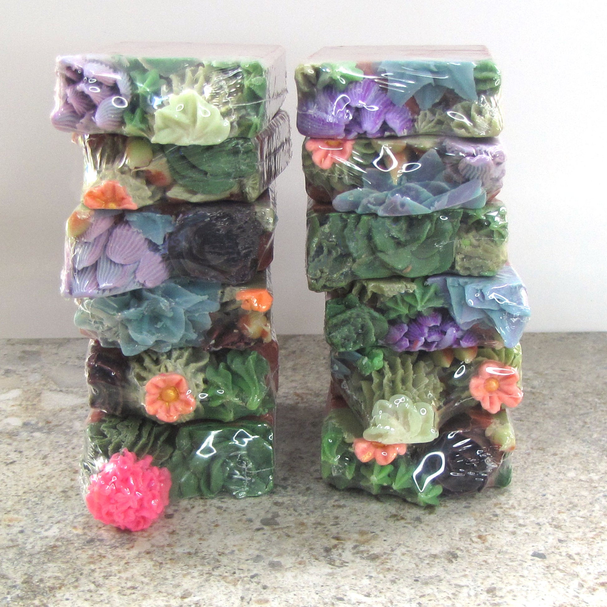 Two stacks of colorful soap bars with soap succulent embeds on top in clear packaging on a neutral background