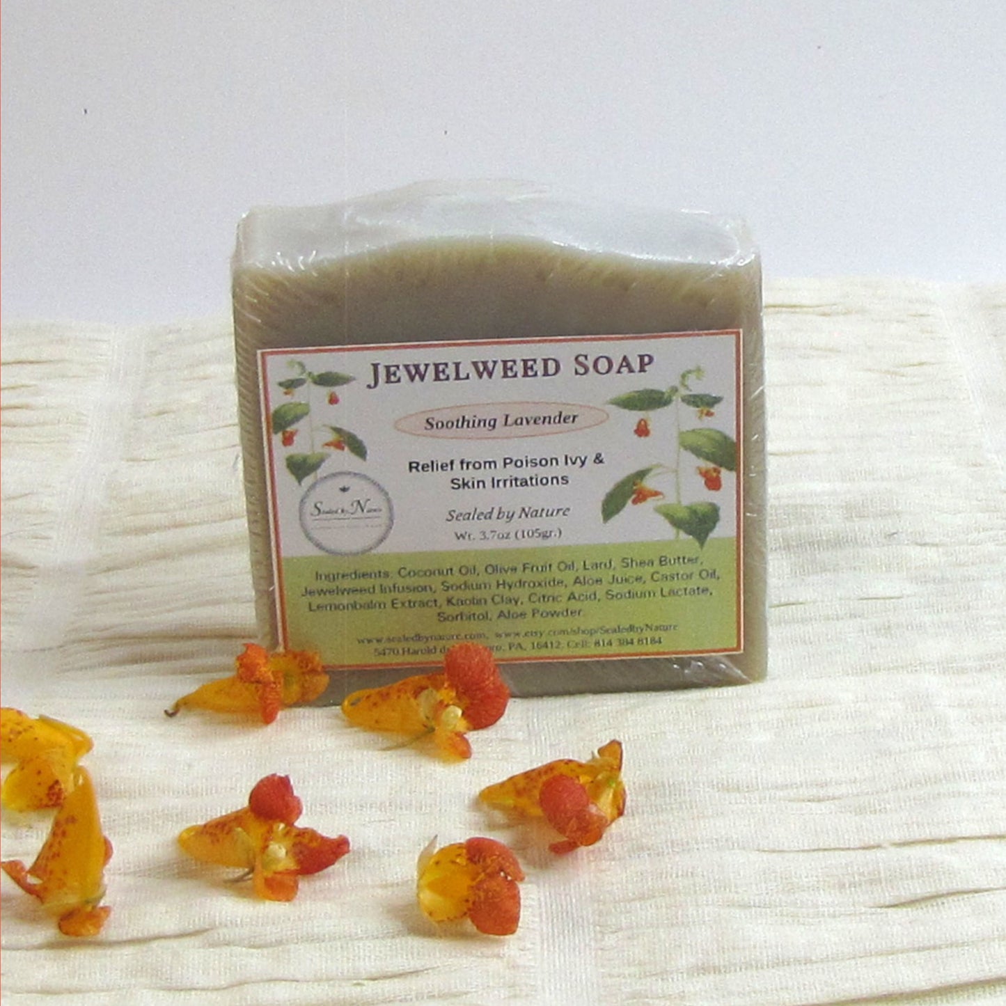 Jewelweed soap bar with packaging on a textured white surface, surrounded by jewelweed flowers.