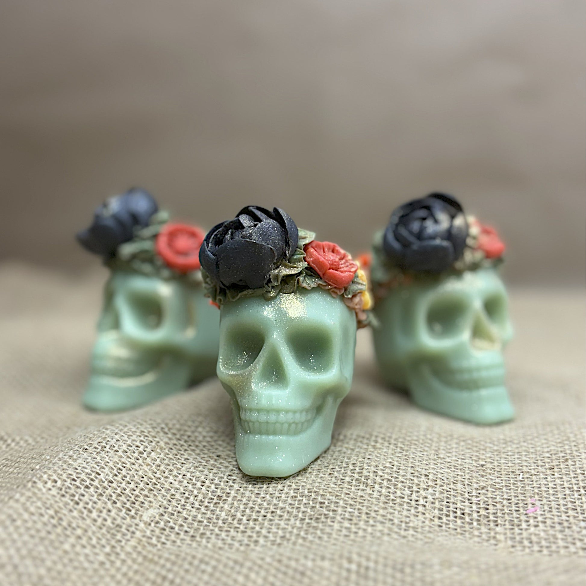 Three green skull-shaped candles with floral arrangements on a textured beige surface.