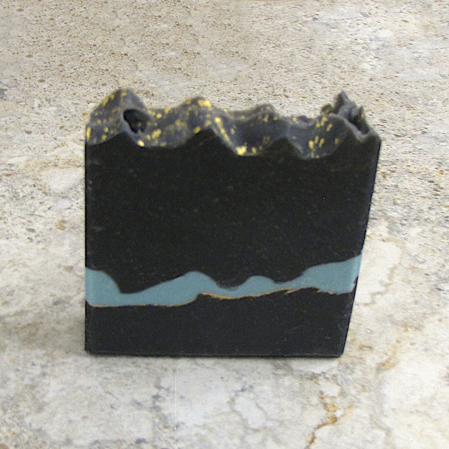 Black soap bar with gold and blue accents on a light stone background