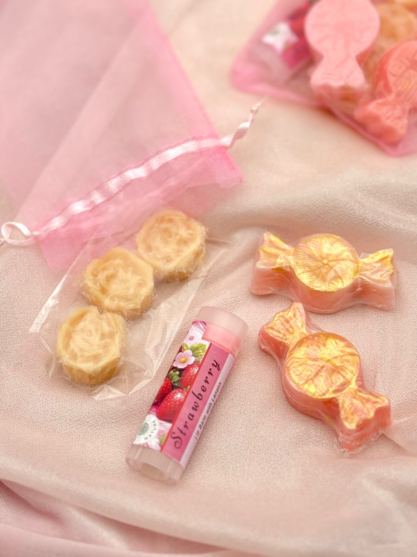 Strawberry-flavored lip balm with candy shaped soaps and three flower shaped solid moisturizers on a pink fabric background
