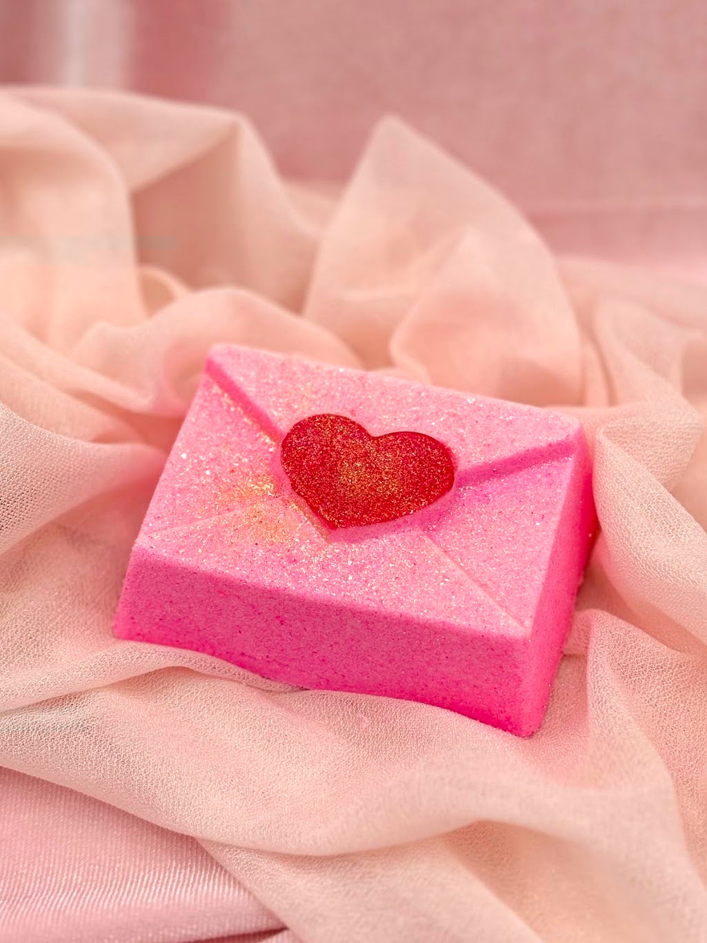 Pink bath bomb shaped like a letter with a red heart on a pink fabric background