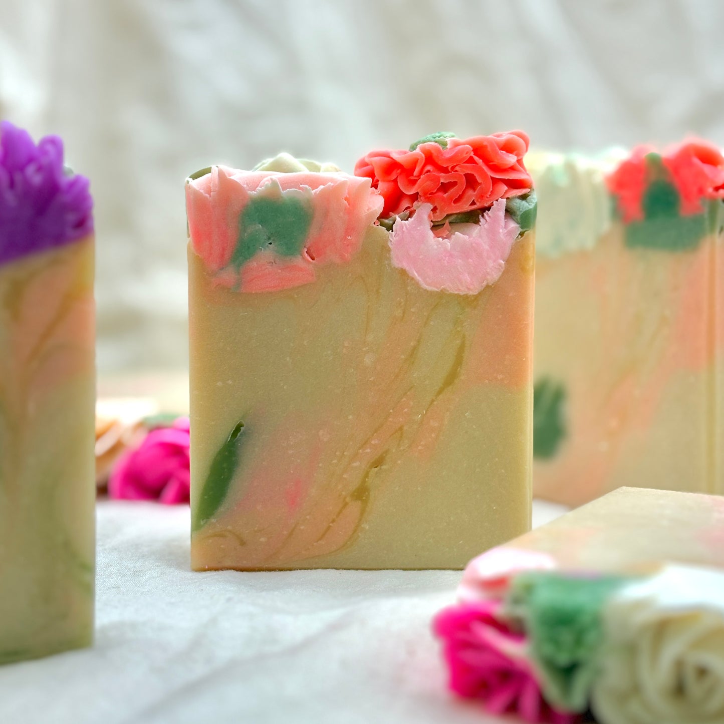 Handcrafted marbled soap bars decorated with sculpted flower tops in pink, coral, green, and white, arranged on a soft white fabric background.