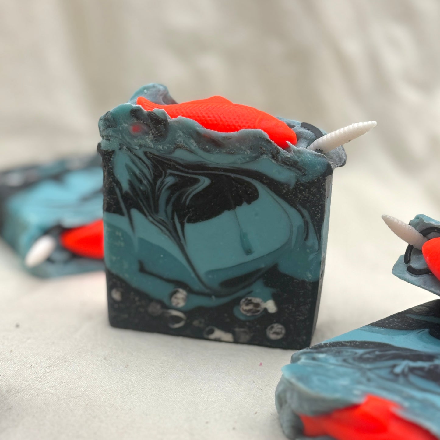 A handmade rectangular soap bar with a swirled black and teal pattern, featuring a bright orange horn-like accent on top and white horn details, shown on a light fabric background.