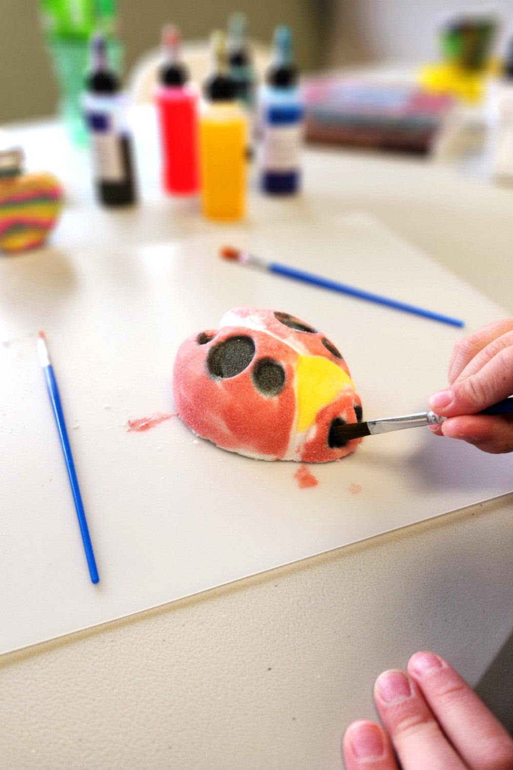 Person painting a ladybug bath bomb with various skin safe art supplies on a table.