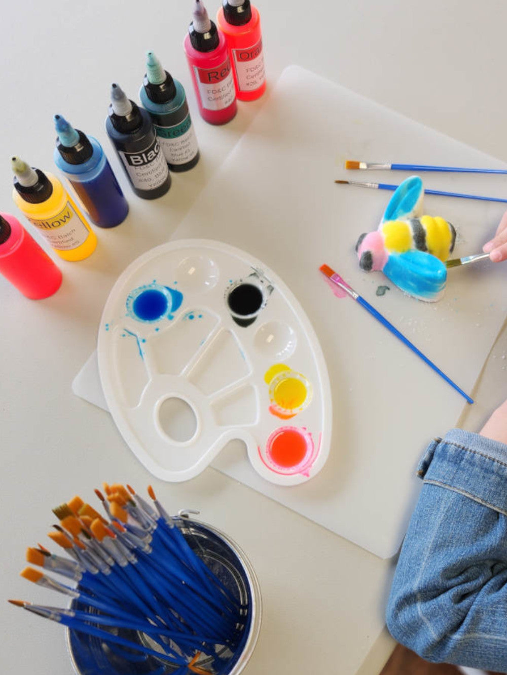 Skin safe art supplies including colorants, a palette with colors, brushes, and a partially painted bee bath bomb on a table.