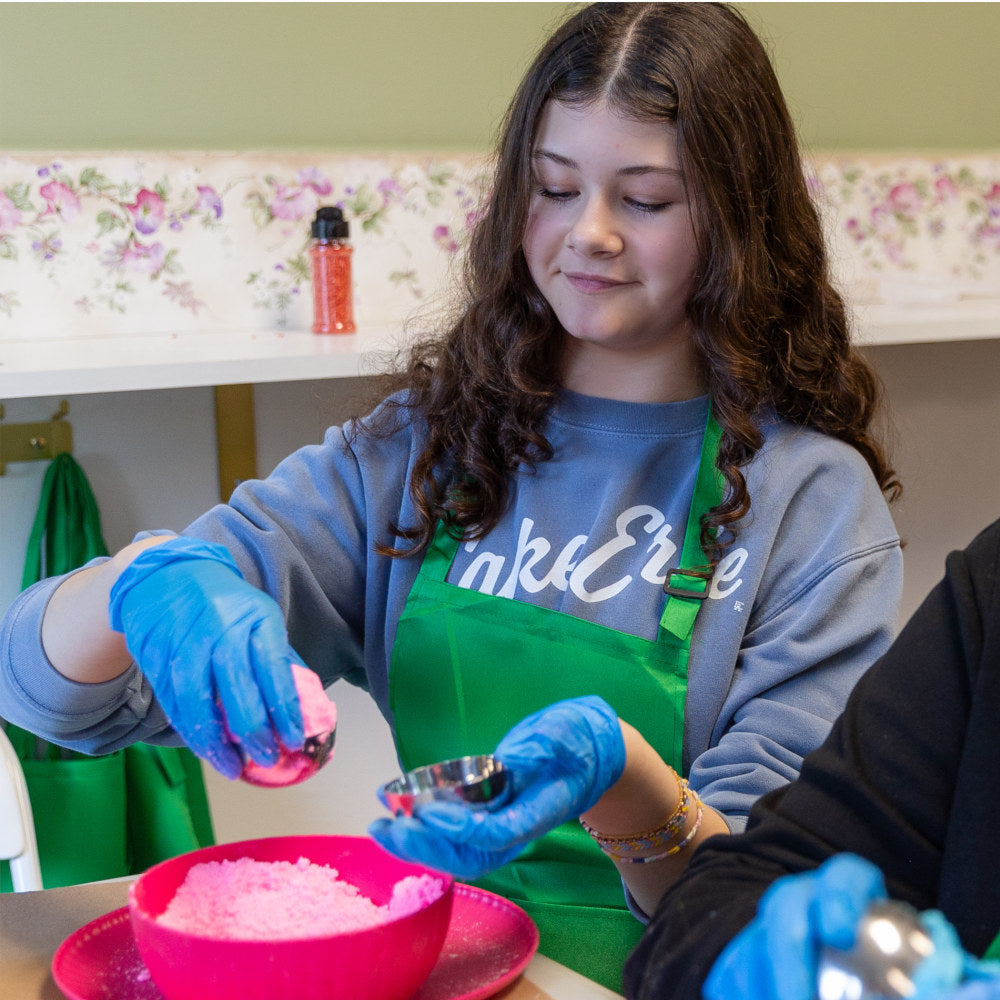 A person wearing a green apron and blue gloves is making a DIY bath bomb.