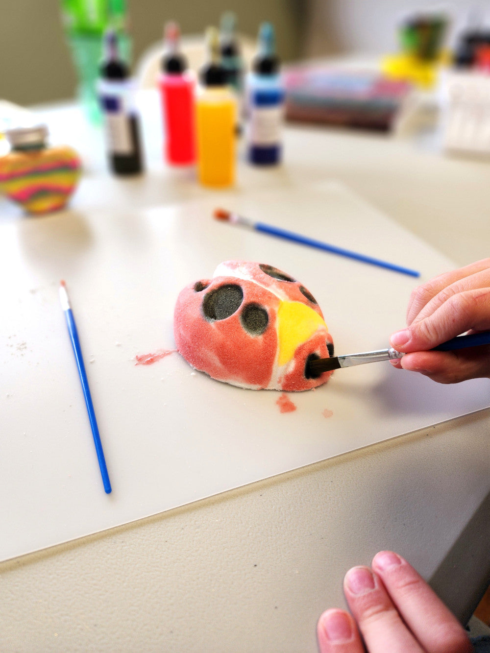 Person painting a small ladybug bath bomb on a table with skin safe art supplies in the background.