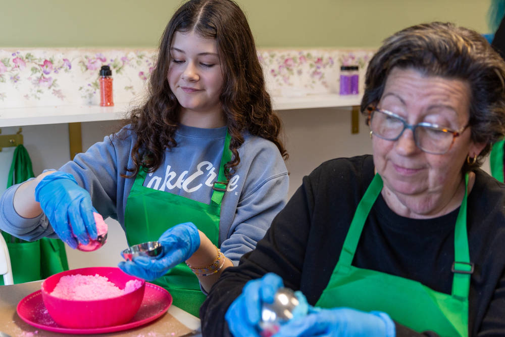 Two people, one adult and one teenager, wearing green aprons and gloves, making bath bombs.