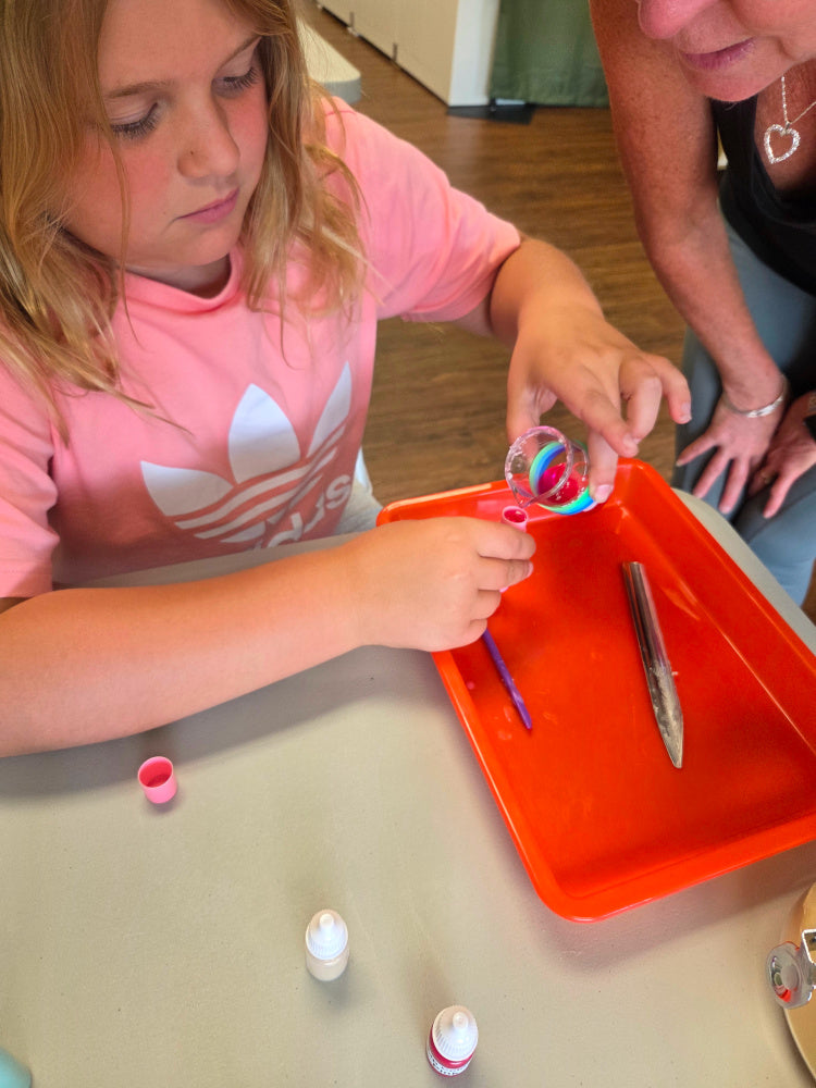 Child and adult engaged in pouring lip balm into a tube.