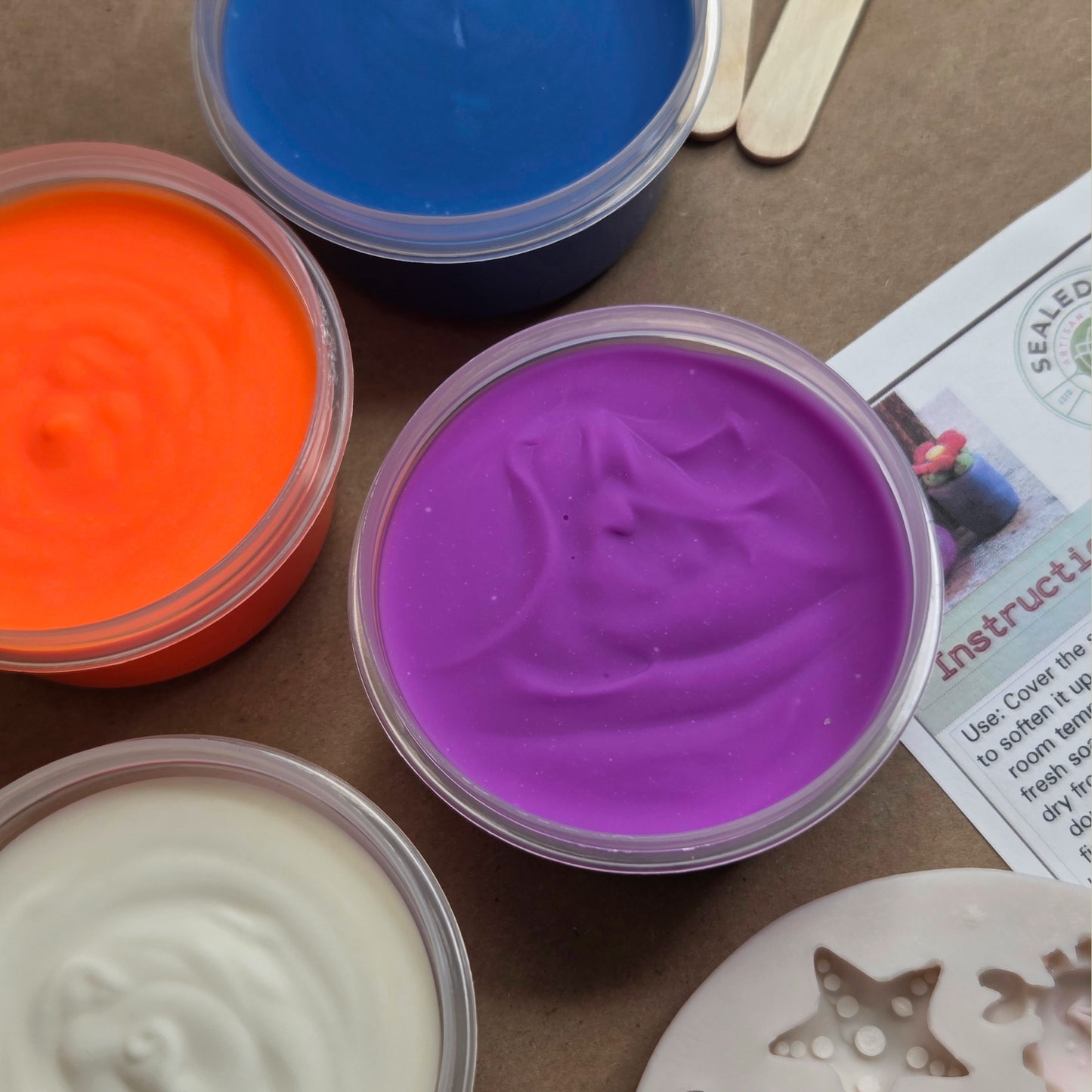 Four containers of colorful slime on a brown surface with a leaflet.