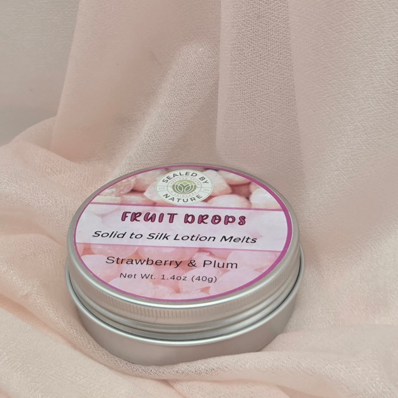 Tin of 'Fruit Drops' solid to silk lotion melts with strawberry & plum flavor on a soft fabric background.