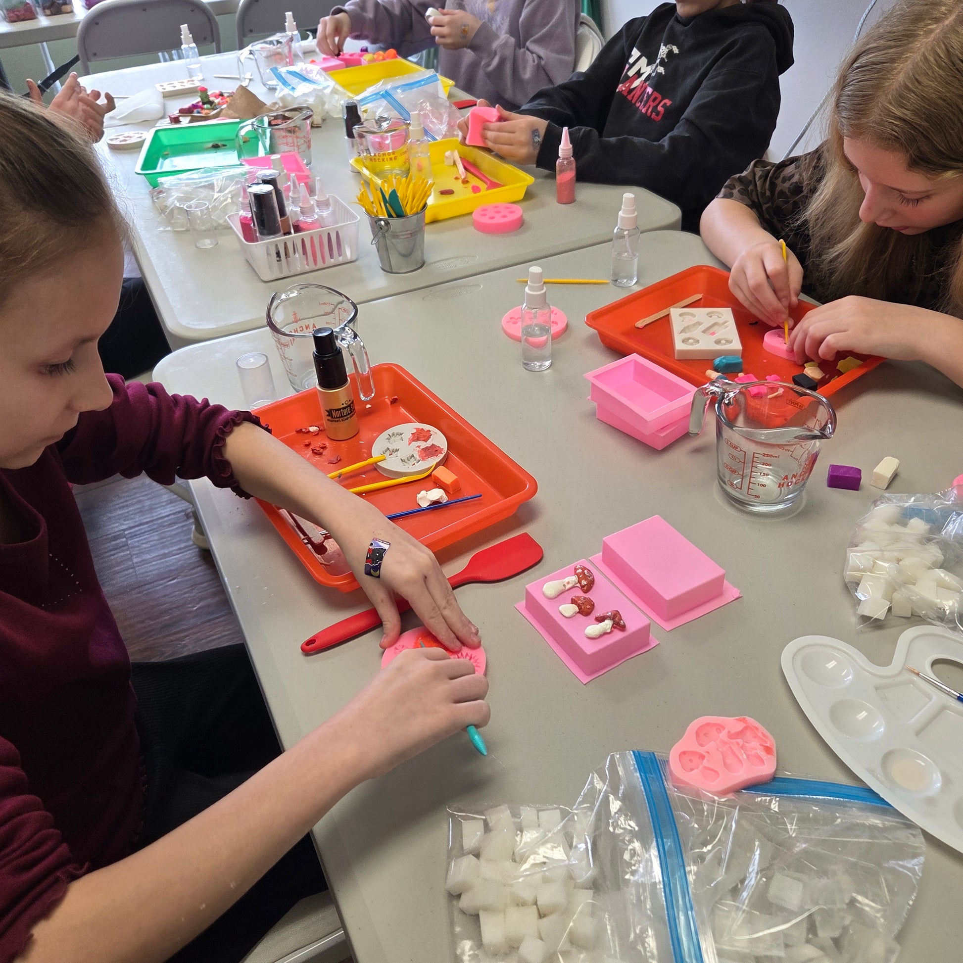 Young girls engaged in a soap making activity with various materials on a table.