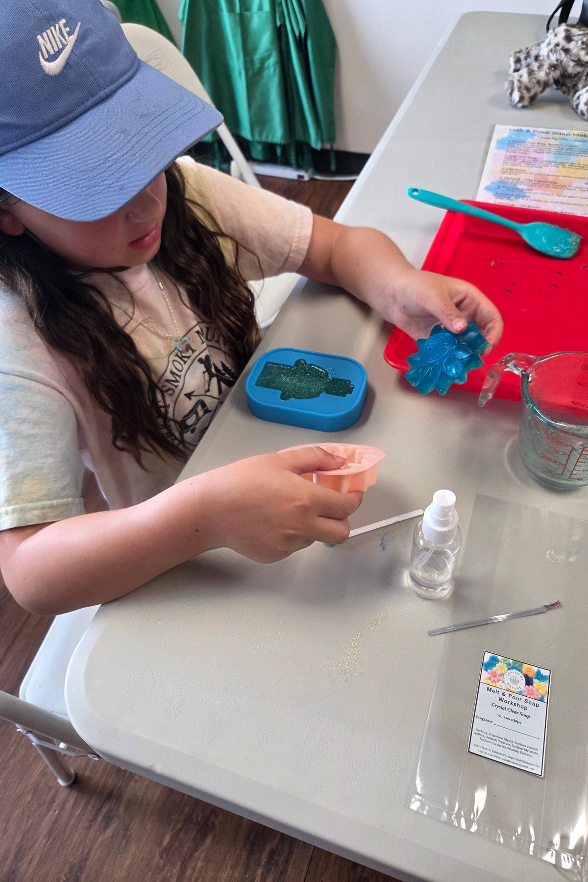 A child wearing a blue cap sits at a gray table, making soap using a mold, a small bottle of liquid in a clear plastic container. A red tray with a turquoise spatula and a blue mold case are nearby.