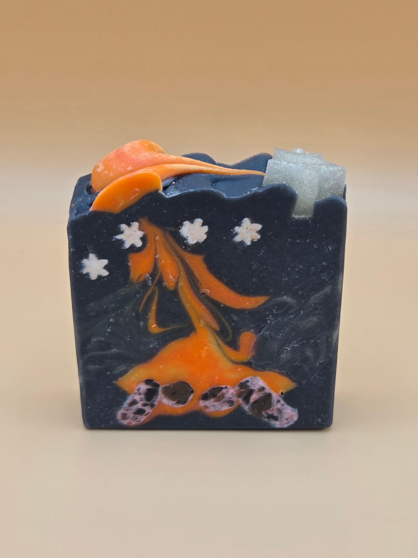 A rectangular bar of soap with a dark navy blue base, featuring an orange fish design, white flower motifs, and a small purple patterned accent, photographed against a plain beige background.