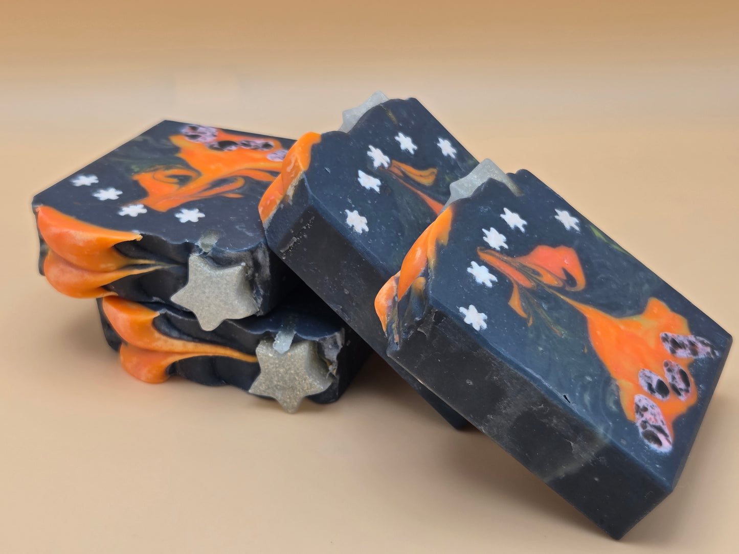 Three handmade soap bars stacked and leaning on a beige background. The soaps are dark navy blue with orange swirls, white star details, and silver glitter accents.
