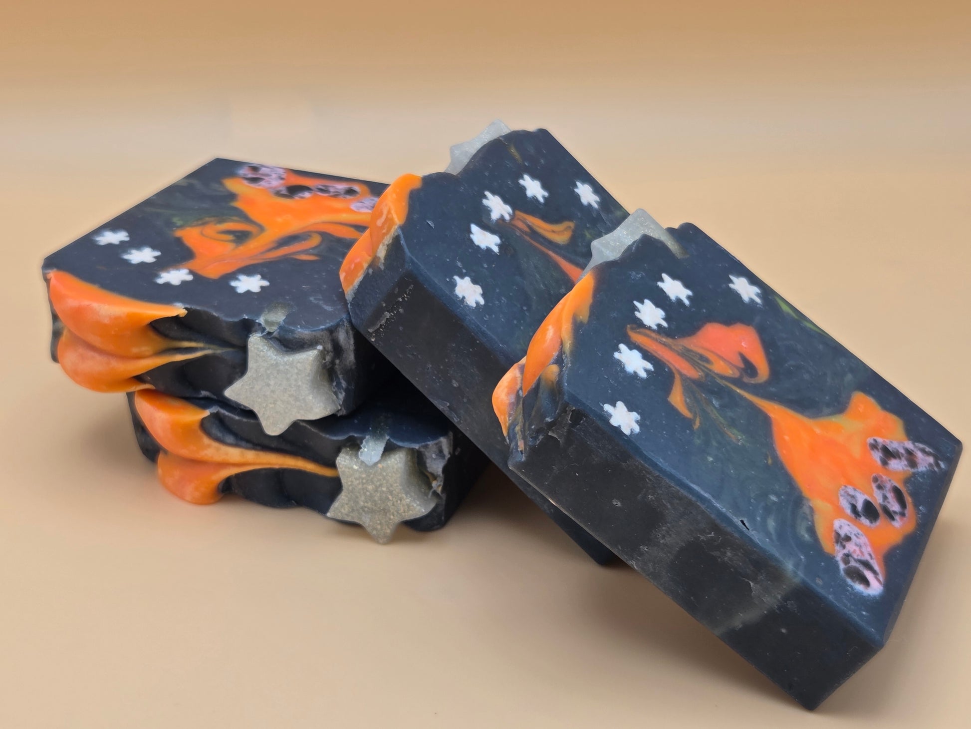 Three handmade soap bars stacked and leaning on a beige background. The soaps are dark navy blue with orange swirls, white star details, and silver glitter accents.