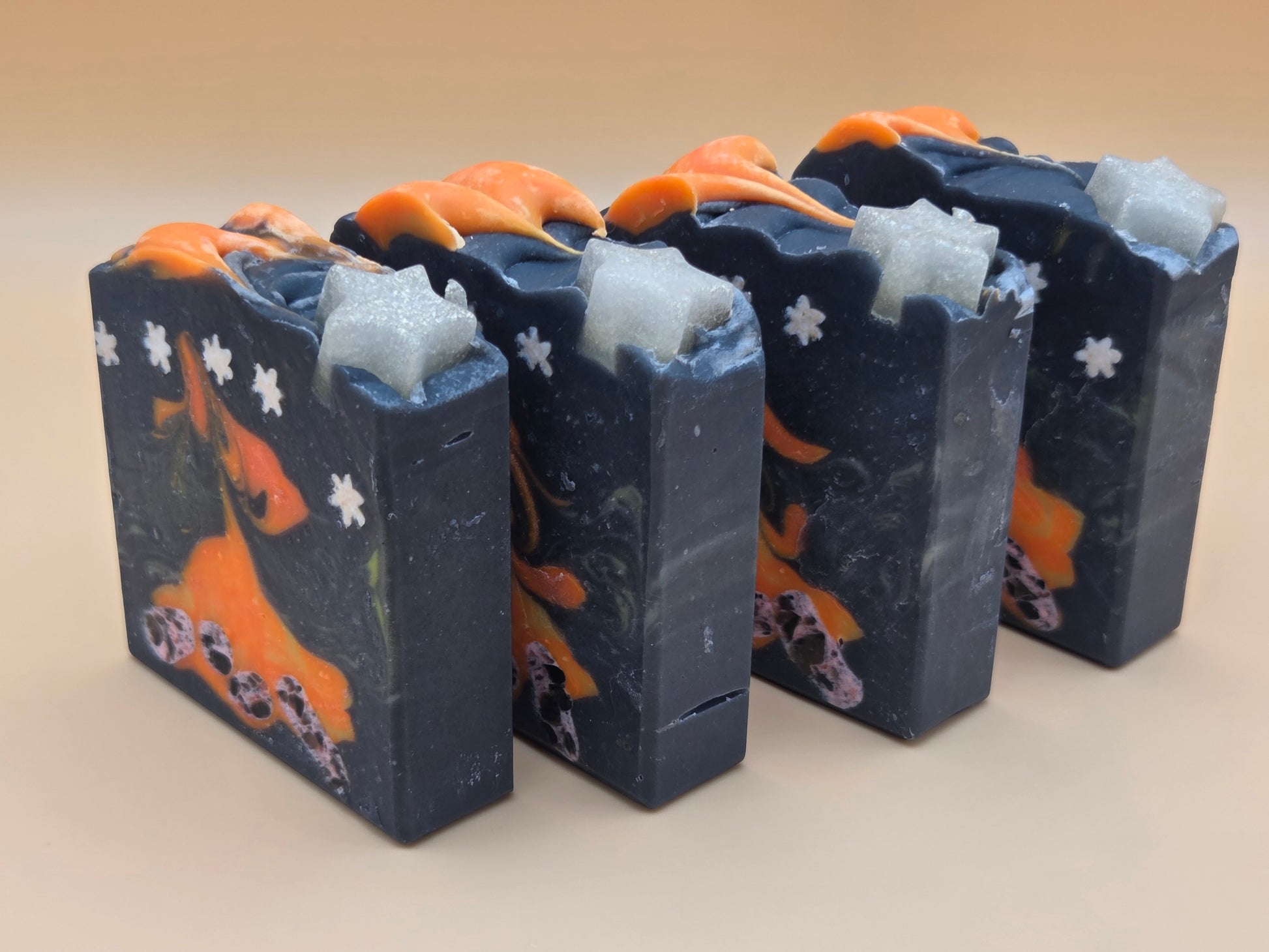 Four handmade rectangular bars of soap standing upright in a row. The soap has a dark navy blue base with orange abstract swirls, small white flower motifs, and light blue-gray textured inclusions near the top, set against a plain beige background.