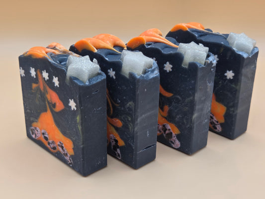 Four handmade rectangular bars of soap standing upright in a row. The soap has a dark navy blue base with orange abstract swirls, small white flower motifs, and light blue-gray textured inclusions near the top, set against a plain beige background.