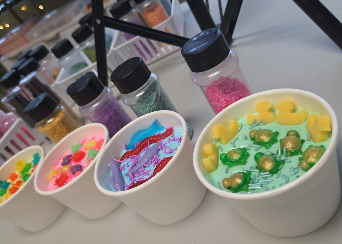 A close-up row of white paper cups filled with colorful sugar scrub and decorated with small soaps - yellow ducks, green and gold turtles, and sprinkles, with small jars of colorful sprinkles behind them on a table.