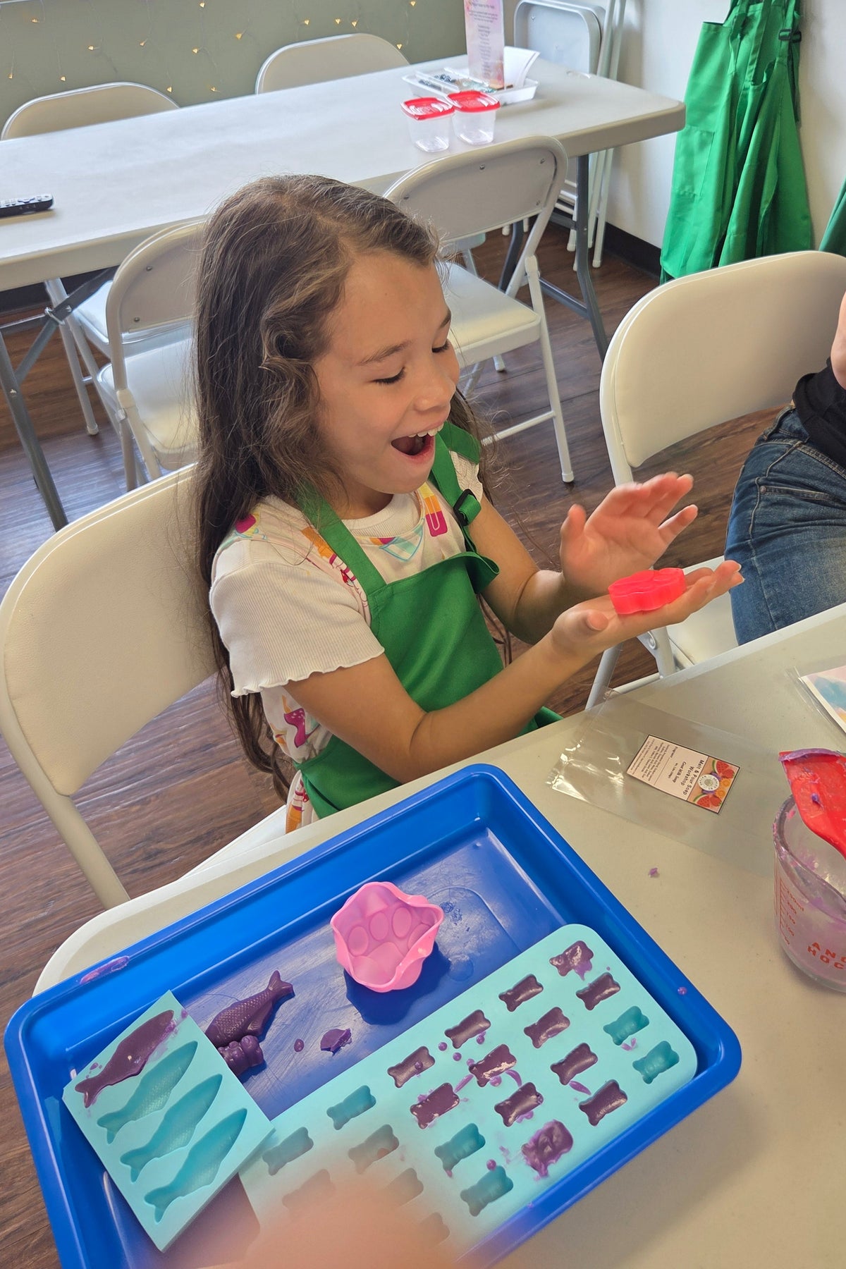 A young girl wearing a green apron smiles while holding a small pink soap at a table, with a blue tray of craft supplies and a red spatula in the foreground. The setting is a workshop interior.