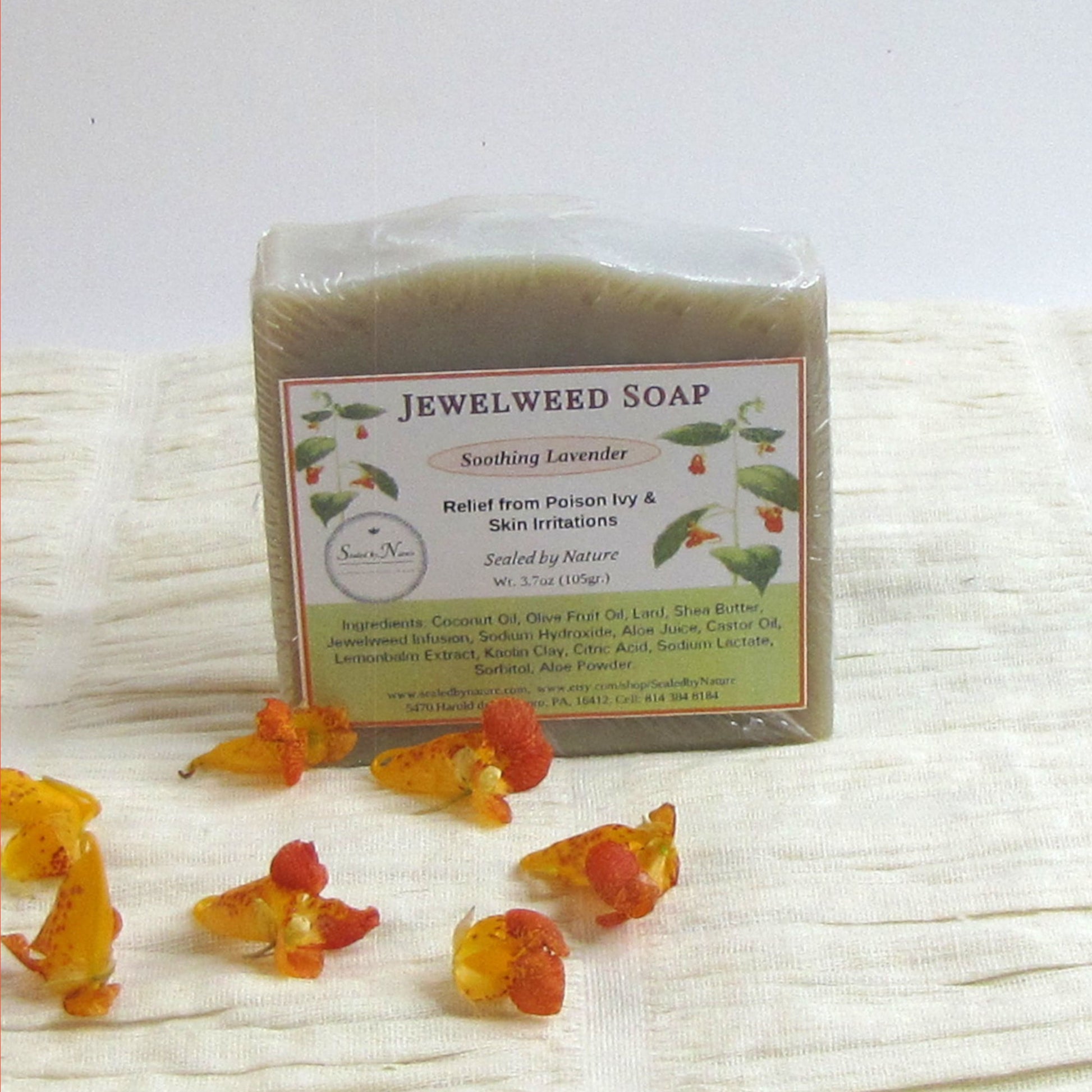 Jewelweed soap bar with packaging on a textured white surface, surrounded by jewelweed flowers.