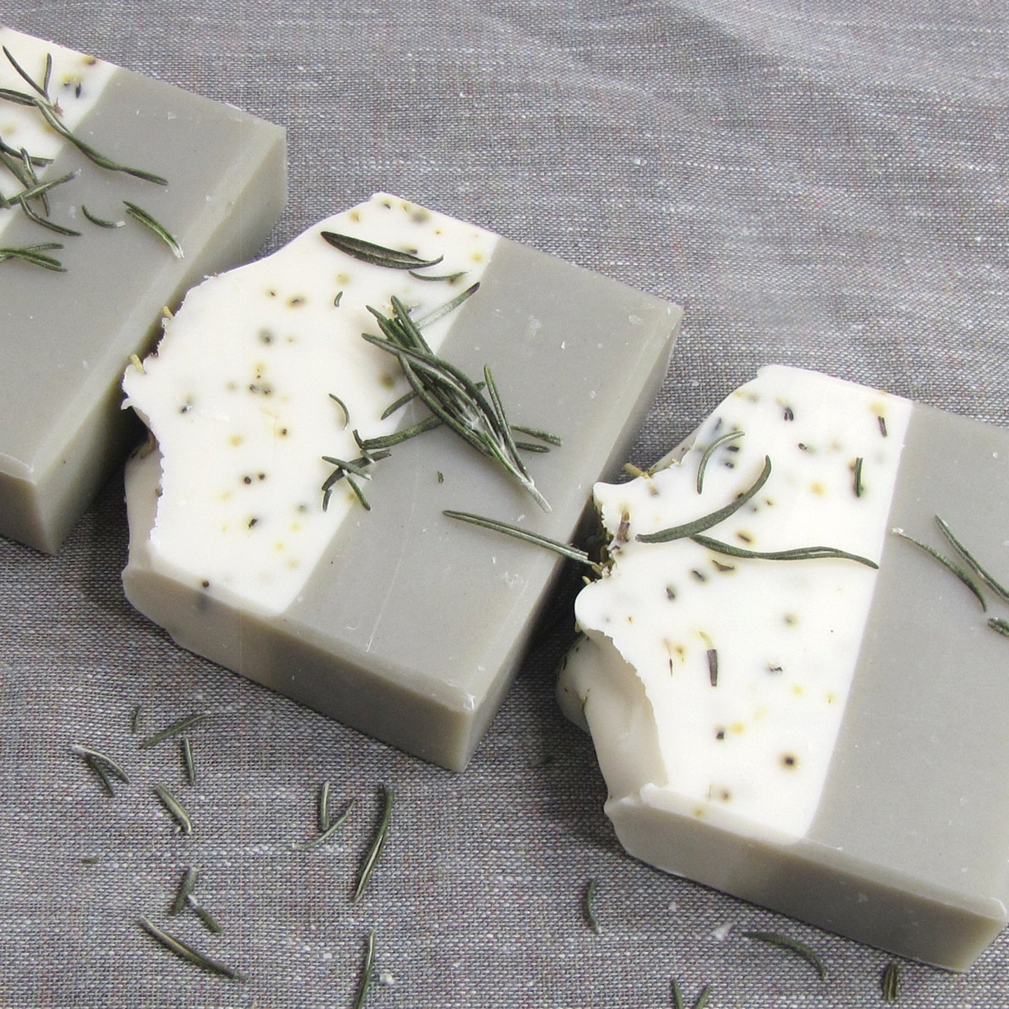 Two layer soap bars with bentonite clay and  rosemary leaves on a gray fabric background