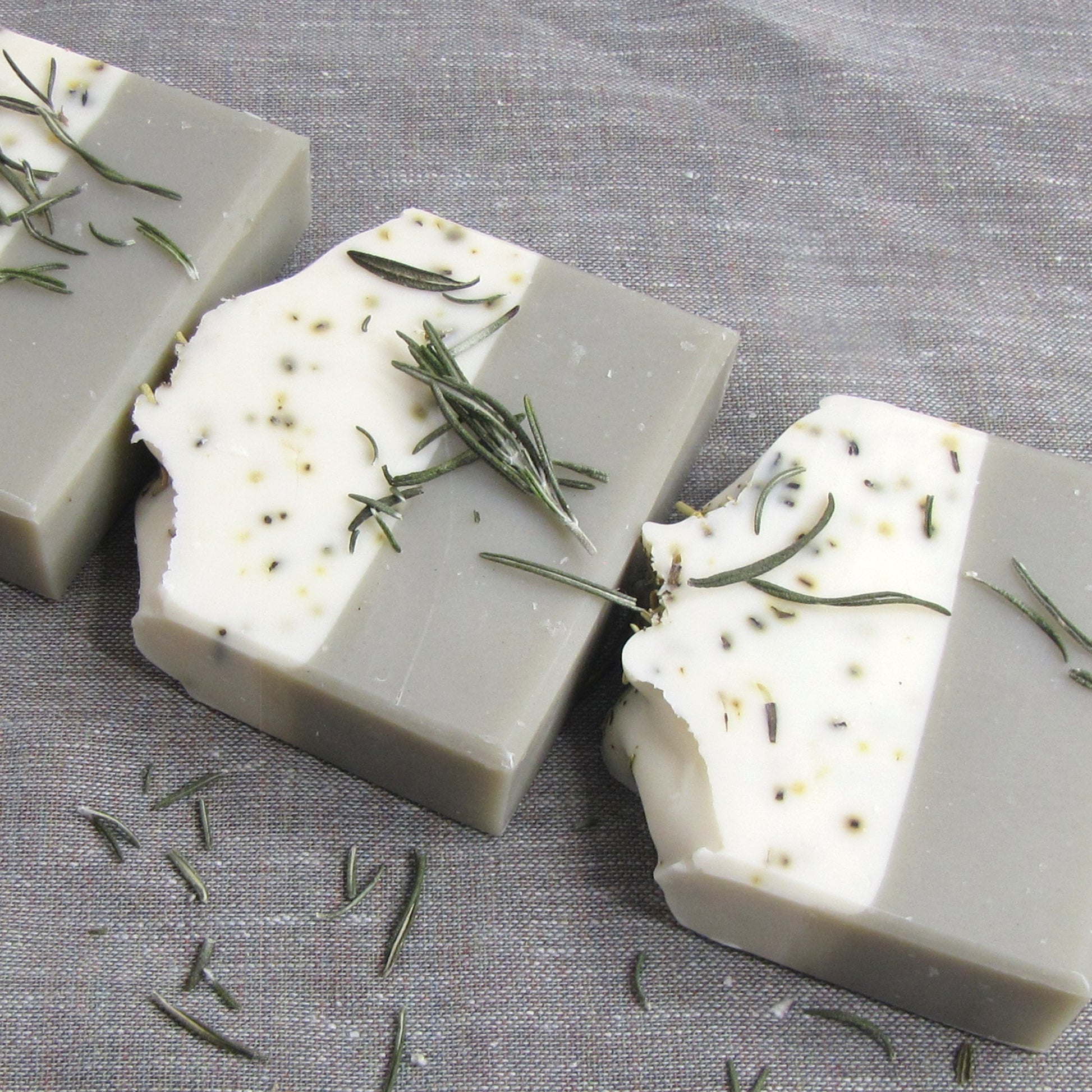 Two layer soap bars with bentonite clay and  rosemary leaves on a gray fabric background