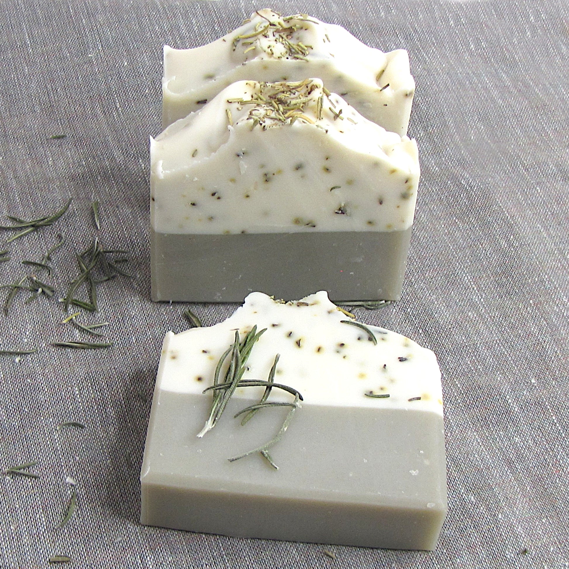 Three bars of soap with rosemary leaves on a gray fabric background