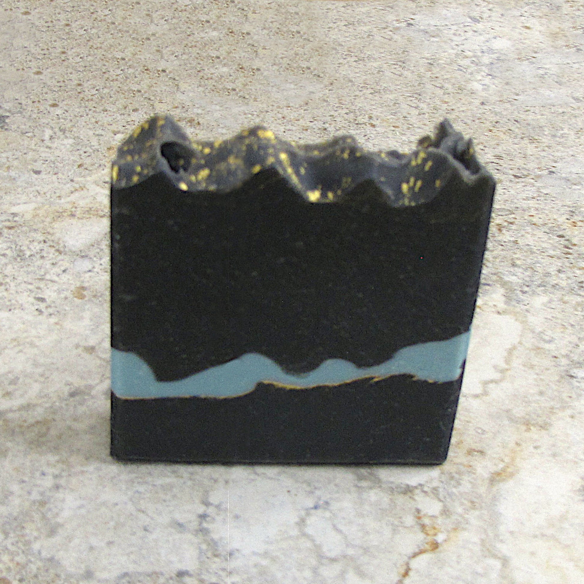 Black soap bar with gold and blue accents on a light stone background