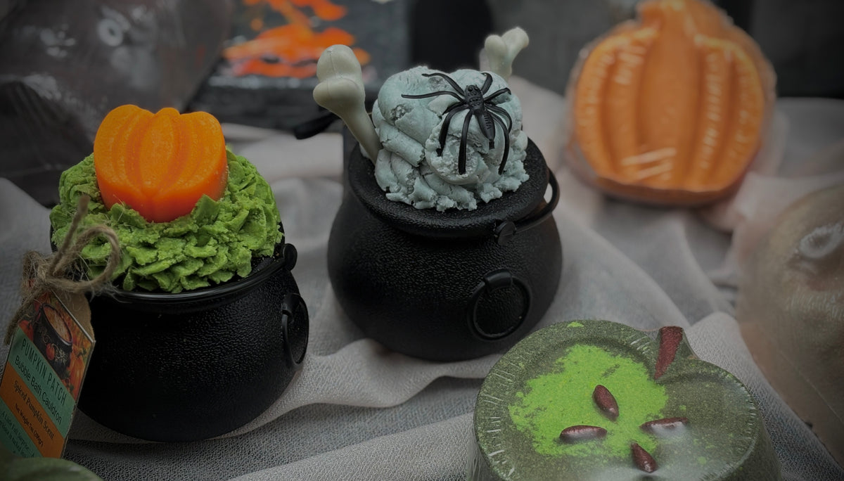 Three Halloween-themed mini bath bombs styled in black cauldrons, with green and white frosting-like tops, orange pumpkin decorations, and a spider ornament, set on a draped fabric surface.