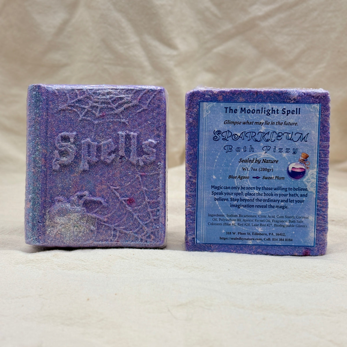 Two lavender-purple rectangular bath bombs in the shape of books standing upright on a light fabric surface. The left bath bomb is embossed with the word 'Spells' and has spiderweb and moon motifs.