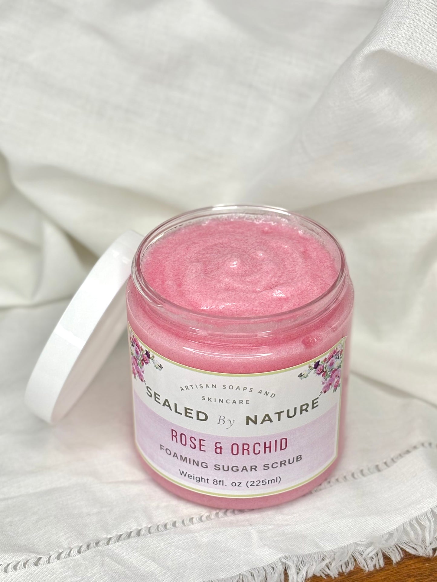 Pink foaming sugar scrub jar with 'Sealed by Nature' label on a white fabric background