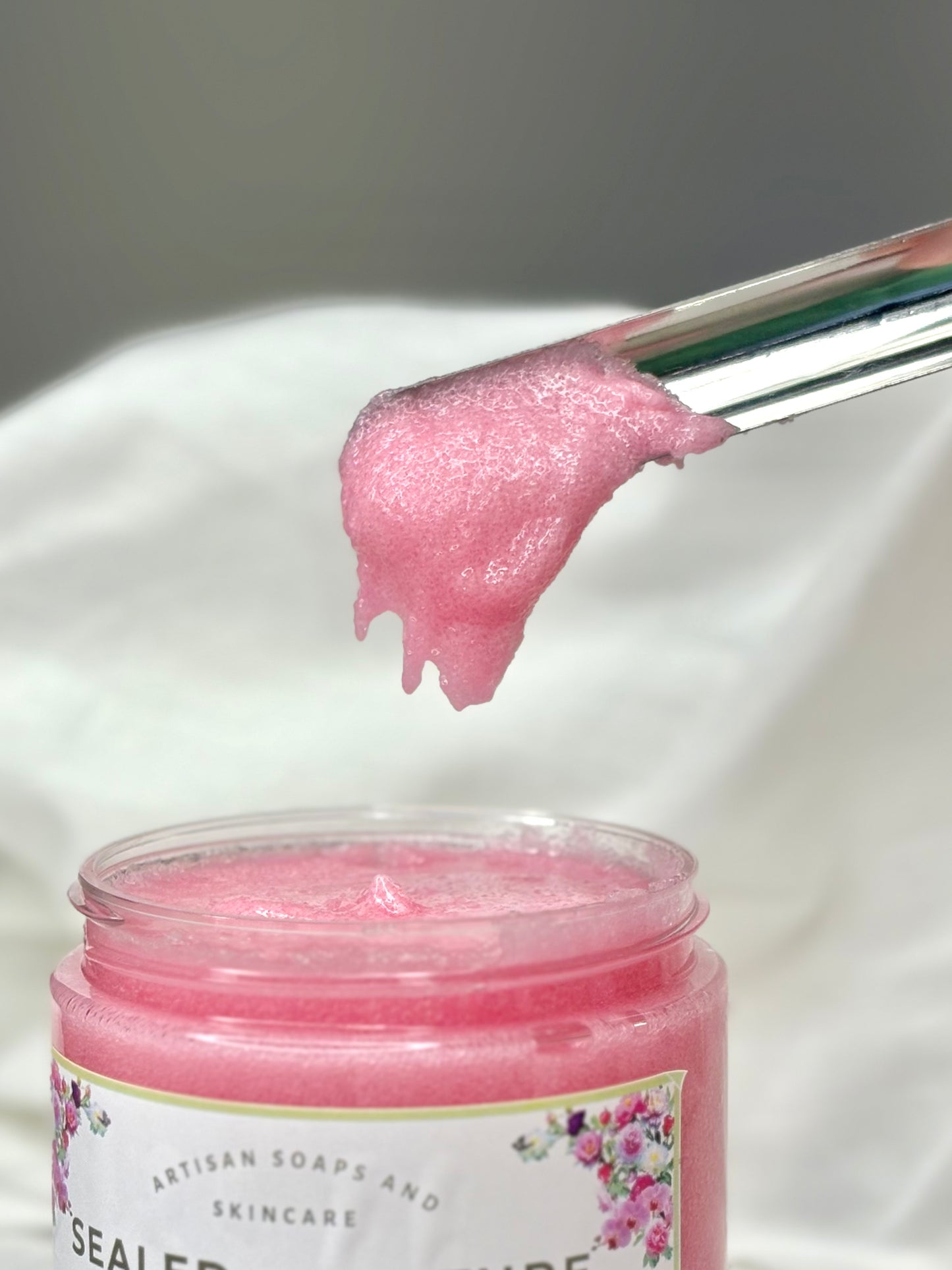 Pink skincare product on a brush with a jar labeled 'Artisan Soaps and Skincare' on a white background