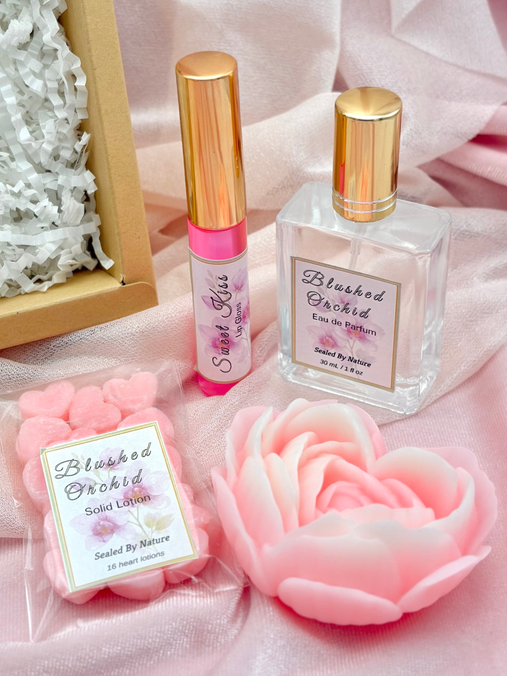 Perfume bottle, solid lotion, lip gloss and soap with 'Blushed Orchid' branding on a pink fabric background.