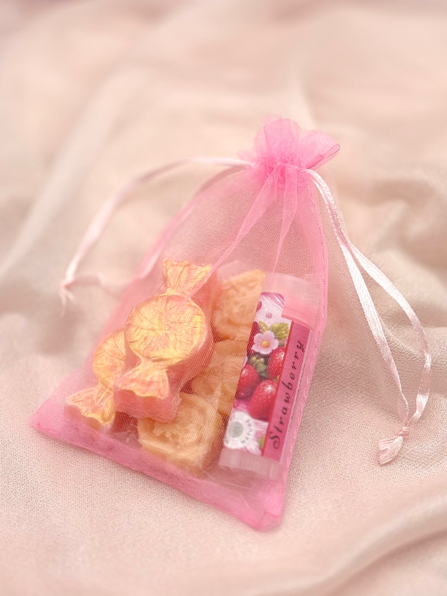 Pink organza bag with strawberry-flavored lip balm, soap candies and a pack of solid moisturizers on a soft pink background