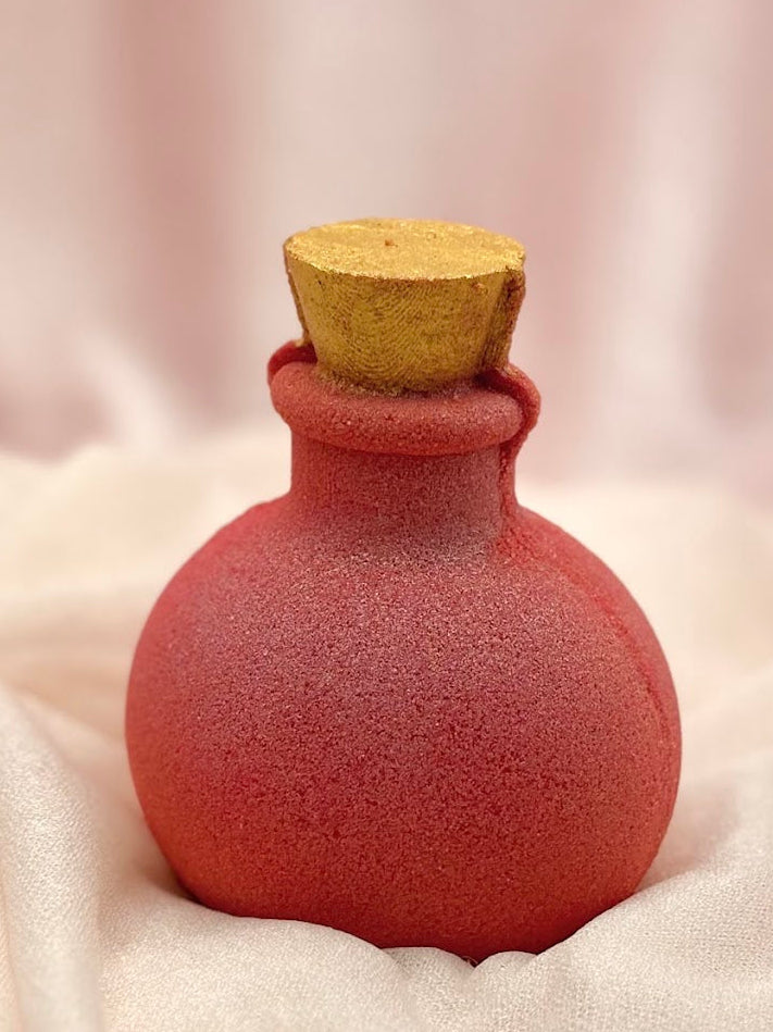 Red potion bottle bath bomb with a gold cork on a soft white fabric background.