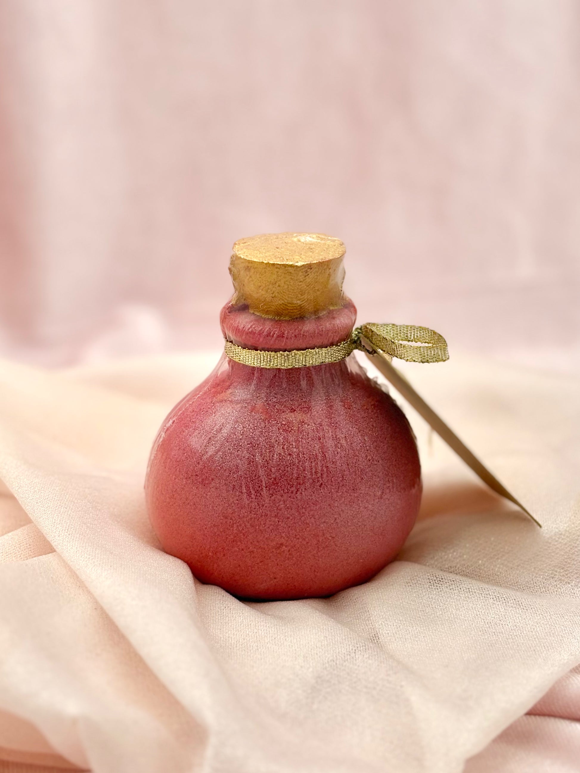 Love Spell Potion Bottle Bath Bomb  with a gold cap and ribbon on a soft pink background.