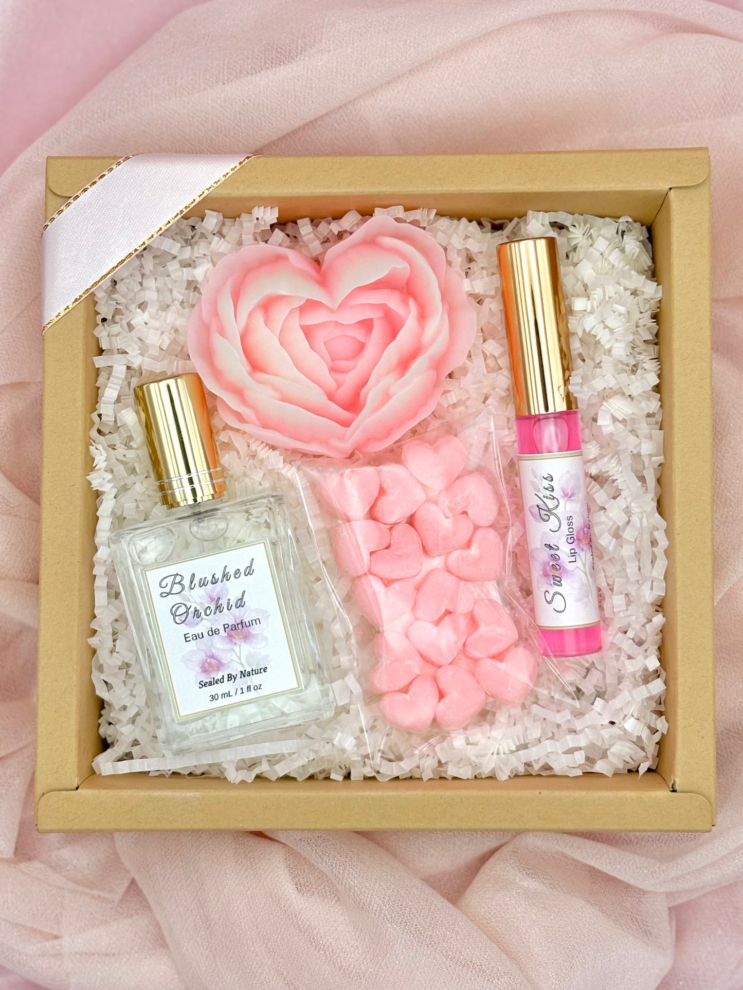 Gift set with perfume, lip gloss, solid heart lotions and soap flower in a decorative box on a pink fabric background.