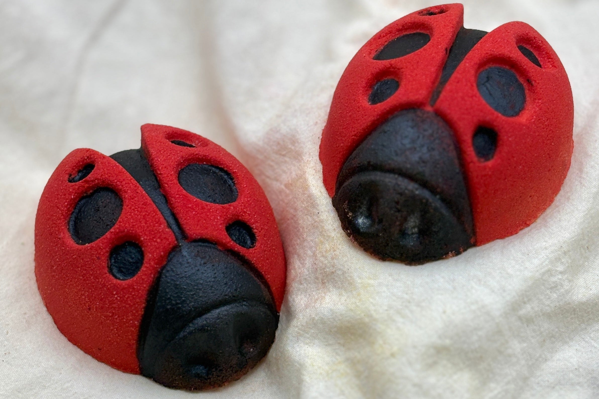 Two red and black ladybug-shaped bath bombs on a white fabric background
