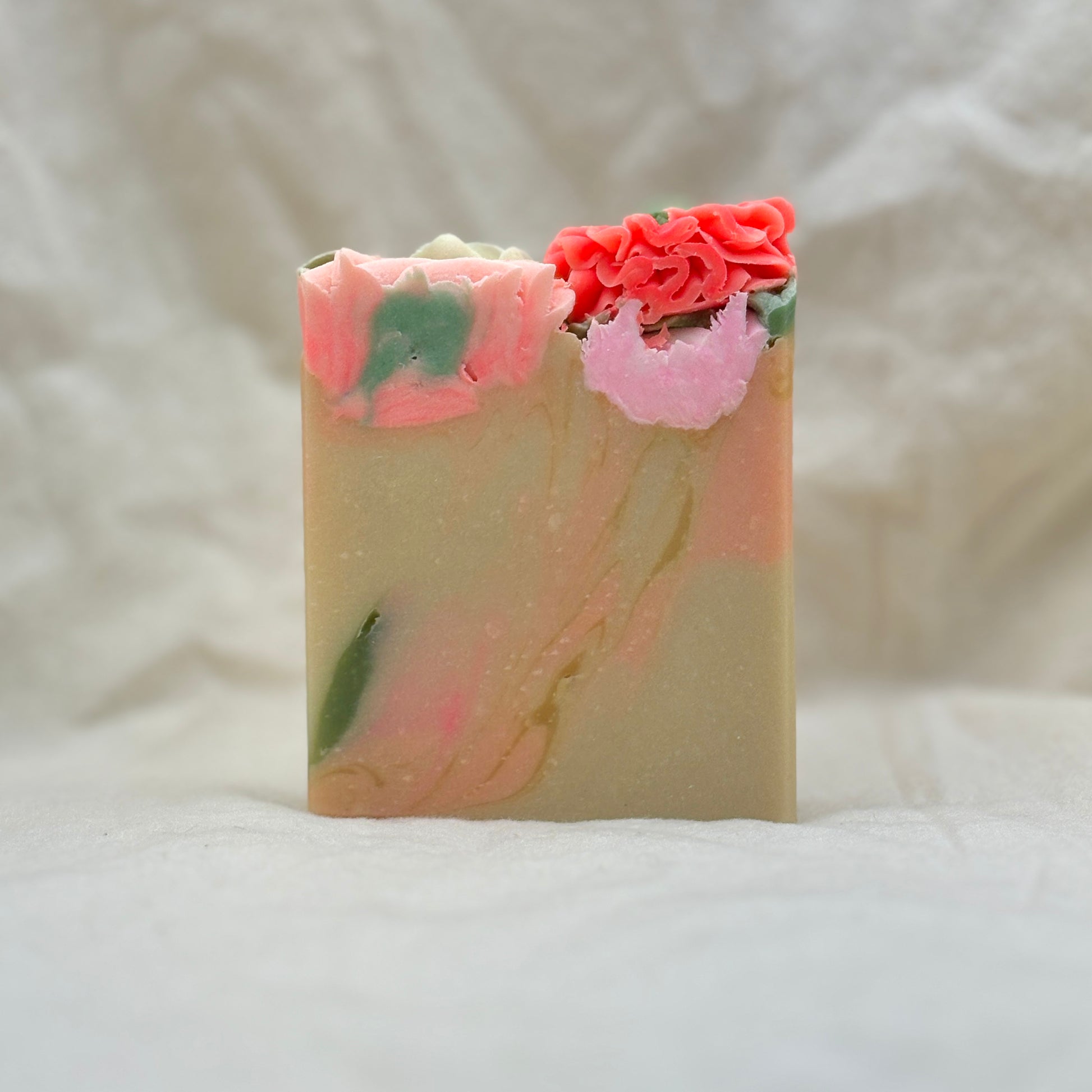 A handmade rectangular bar of soap with a marbled beige base, pink and green swirls, and decorative flower tops in coral pink and light pink, set against a soft white fabric background.