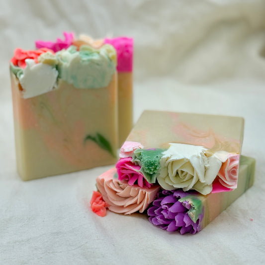 Two decorative handmade soap bars with sculpted flower tops, photographed on a soft white fabric background.