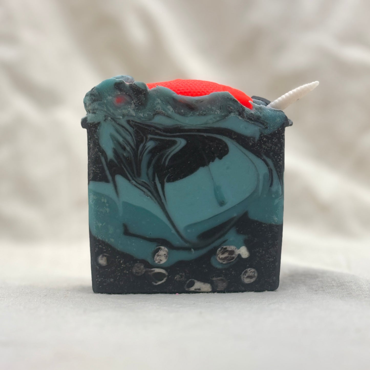A rectangular handmade soap bar with a swirled teal and black marbled pattern, featuring a bright red top section, a small white horn-like protrusion, and embedded clear stones, photographed on a soft light gray fabric background.