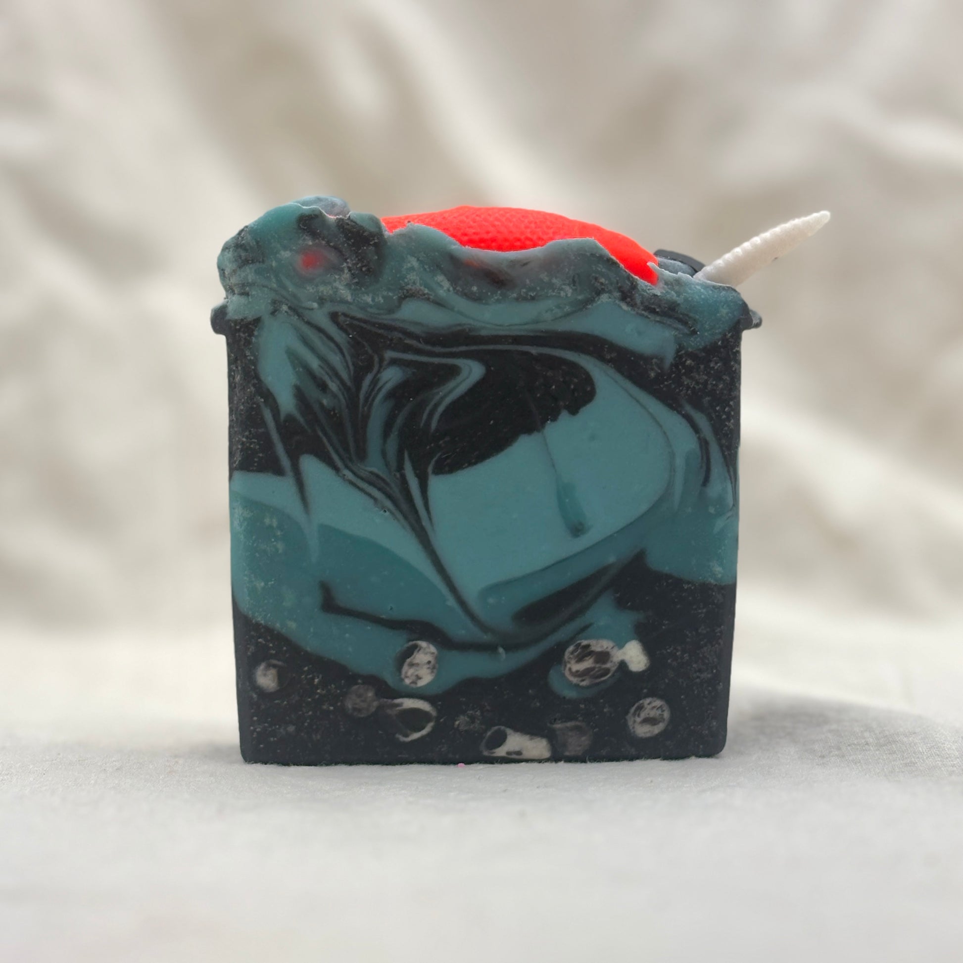A rectangular handmade soap bar with a swirled teal and black marbled pattern, featuring a bright red top section, a small white horn-like protrusion, and embedded clear stones, photographed on a soft light gray fabric background.