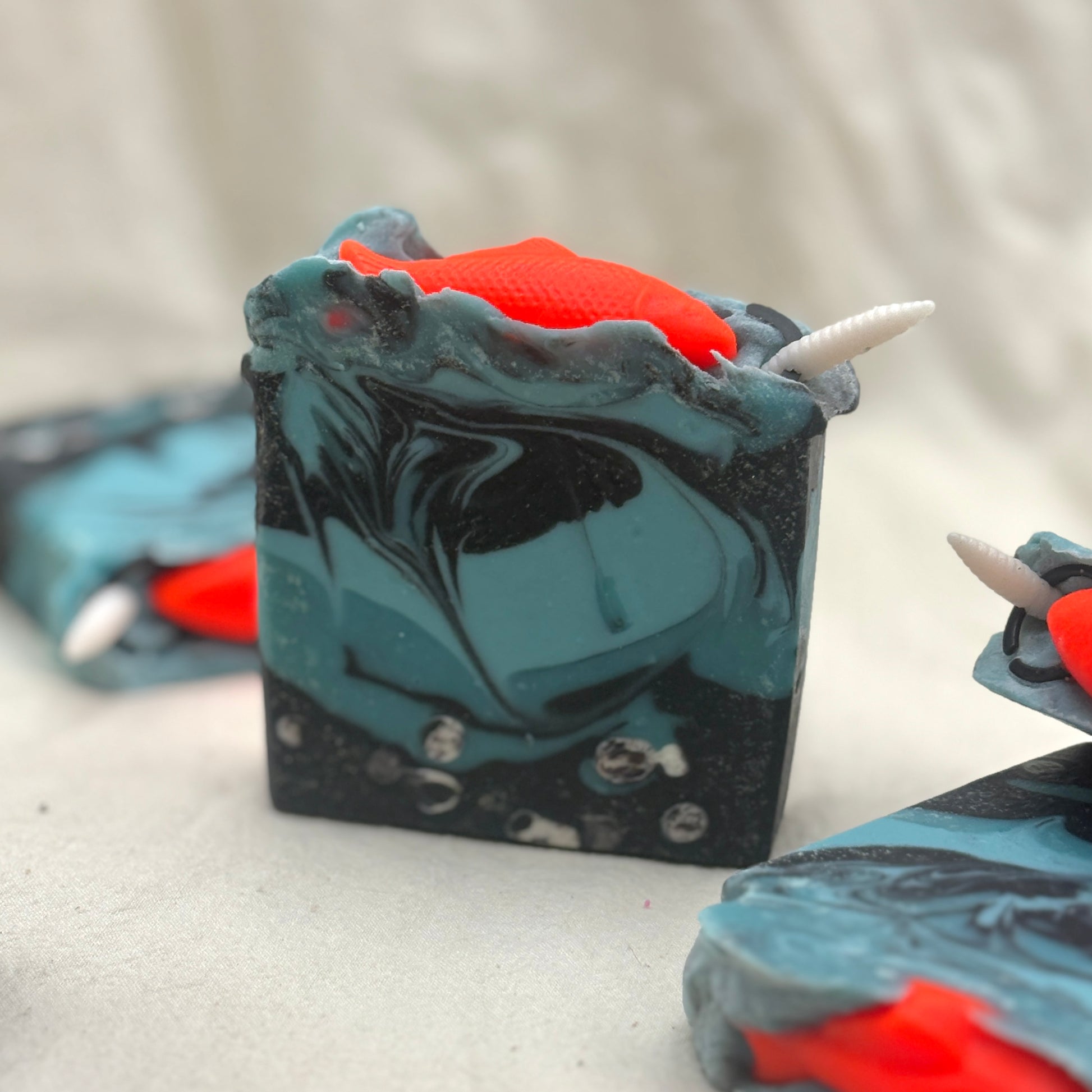 A handmade rectangular soap bar with a swirled black and teal pattern, featuring a bright orange horn-like accent on top and white horn details, shown on a light fabric background.