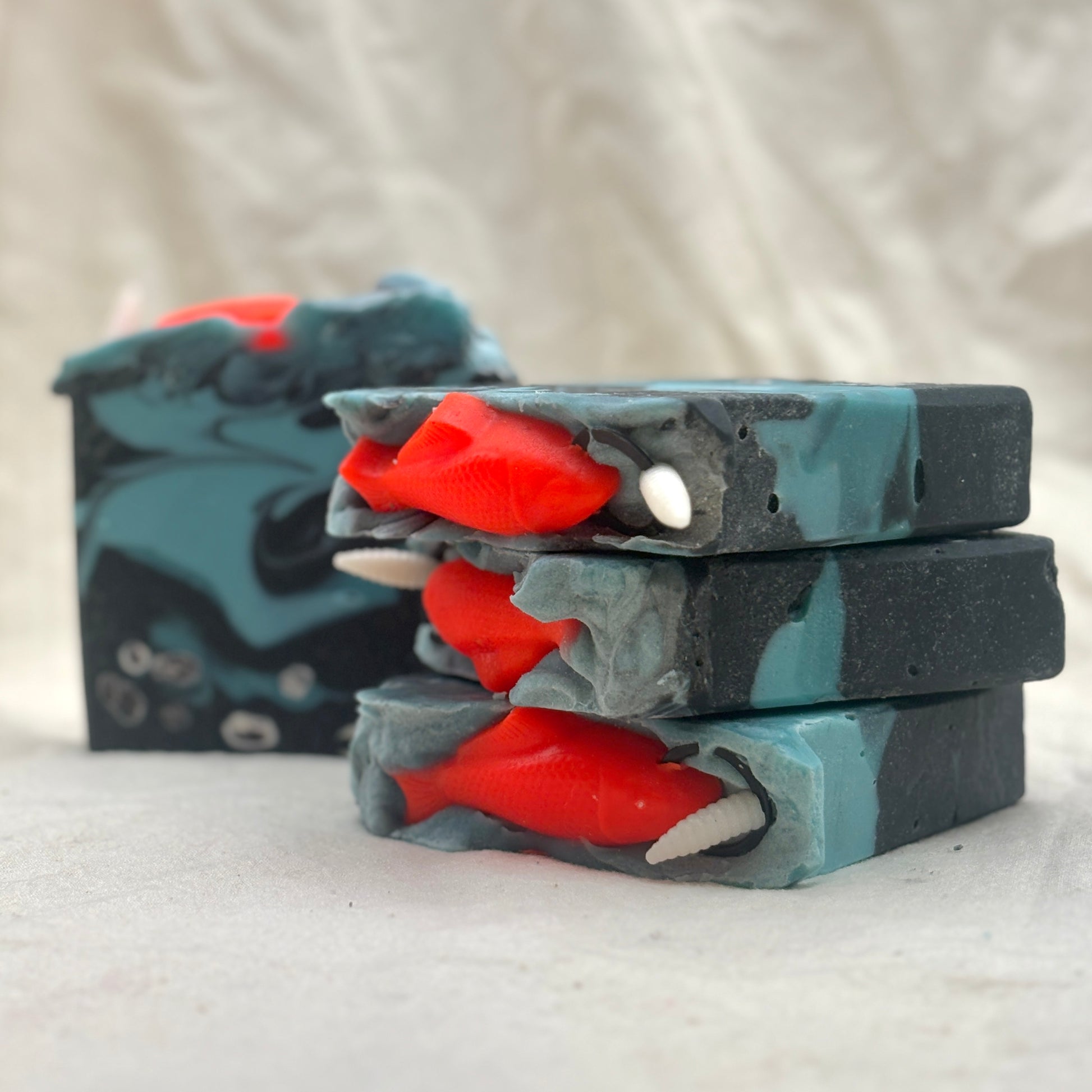 Three stacked rectangular bars of handmade soap with a dark gray and teal swirled pattern, featuring bright red fish-shaped inclusions and small white shell details, photographed on a light fabric background.