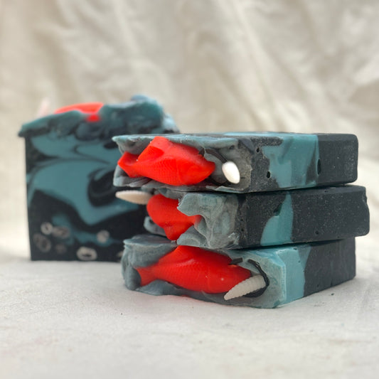 Three stacked rectangular bars of handmade soap with a dark gray and teal swirled pattern, featuring bright red fish-shaped inclusions and small white shell details, photographed on a light fabric background.