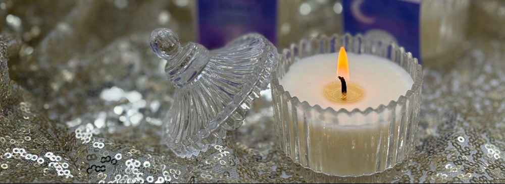 Small lotion candle in a decorative  crystal holder with two product boxes in the background on a glittery surface.