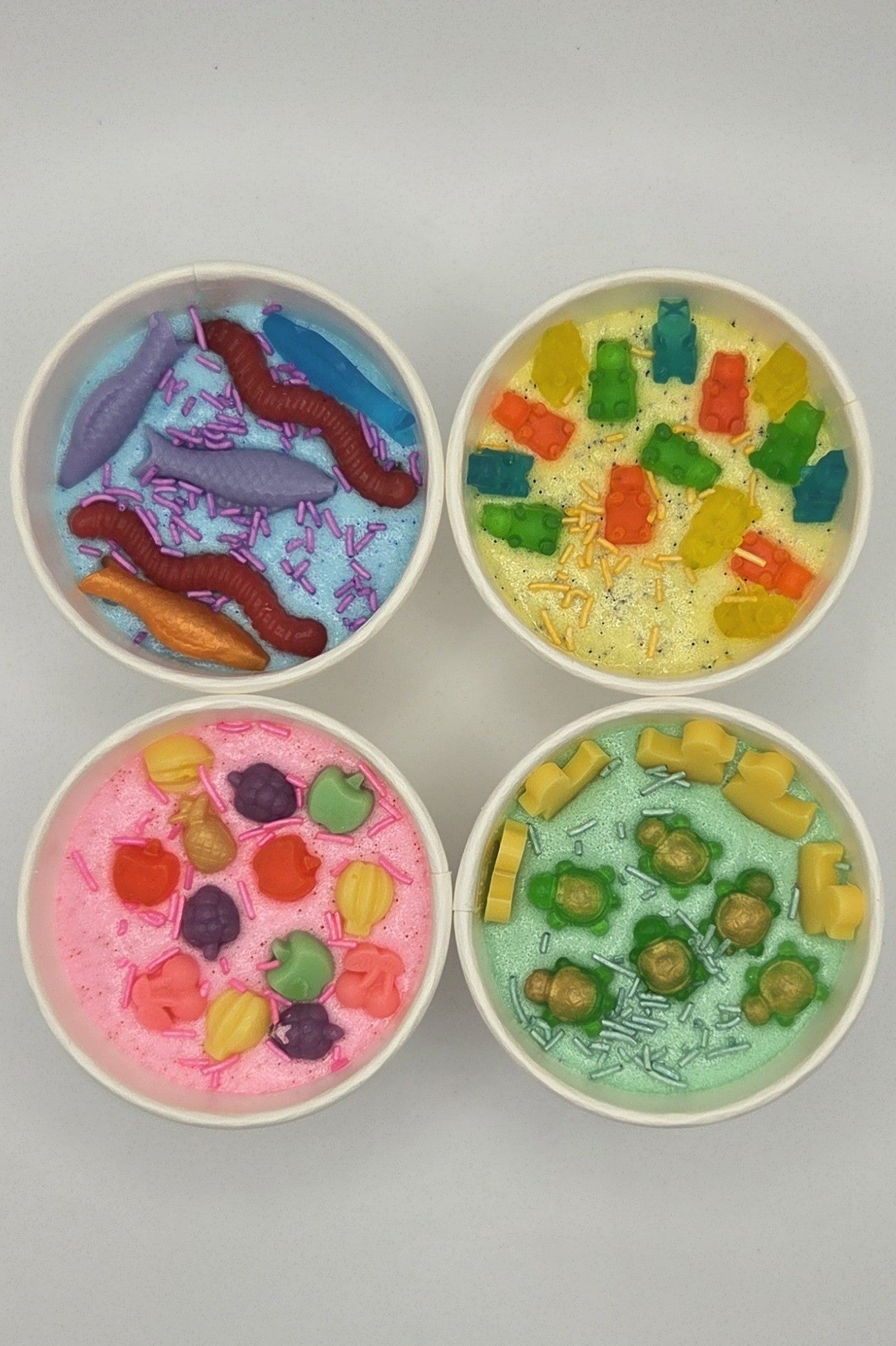 Four small white bowls of sugar scrubs with colorful candy-like toppings. The top-left bowl contains purple, red, and orange pieces on a blue base; the top-right bowl has green, yellow, and orange pieces on a pale yellow base; the bottom-left bowl has pink, orange, and purple pieces on a pink base; the bottom-right bowl has green and yellow pieces on a mint green base.